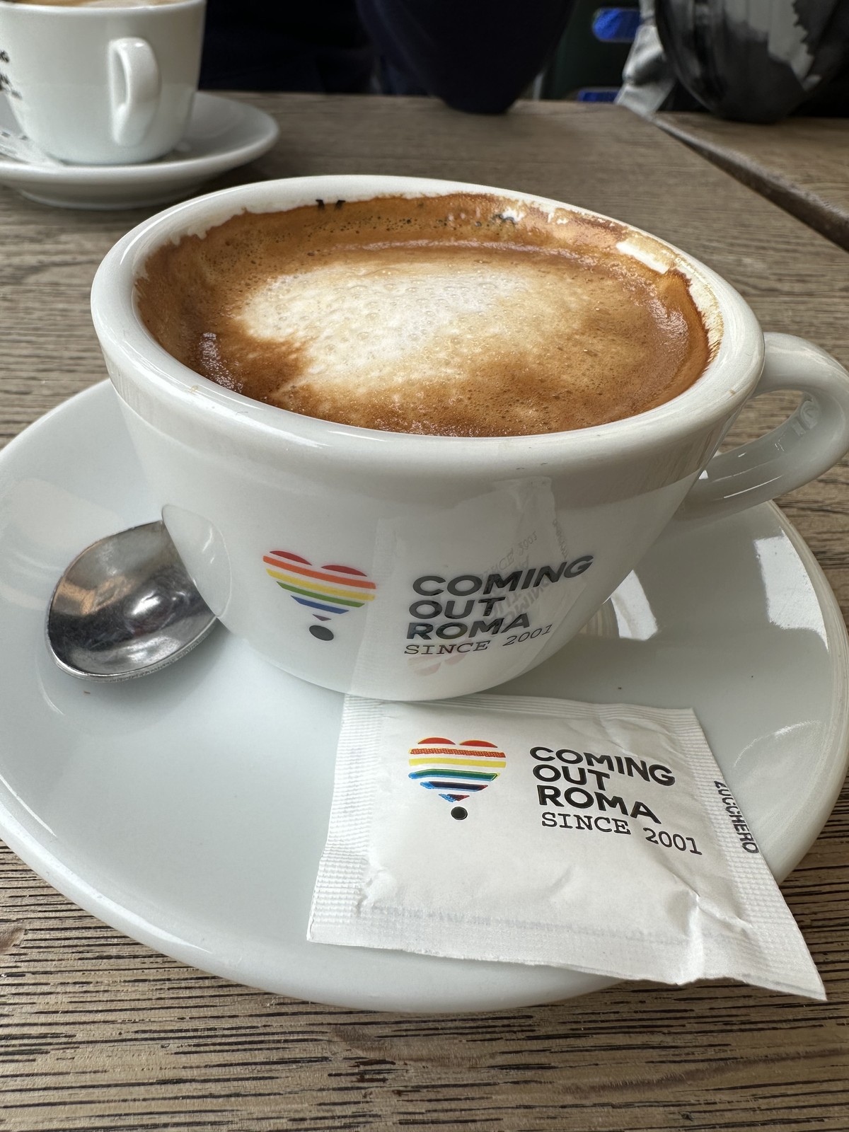 Cappuccino in a Coming Out Roma branded cup with rainbow heart logo, across from the Colosseum
