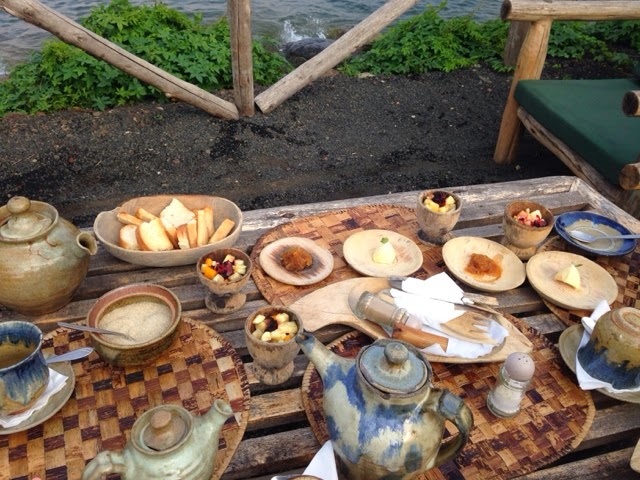 Lakeside breakfast spread with teapots, bread, fruit, and dishes on a rustic wooden table beside Lake Kivu