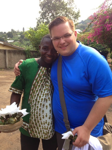 The author in a blue t-shirt posing with a Rwandan friend, both smiling with hills and flowers in the background