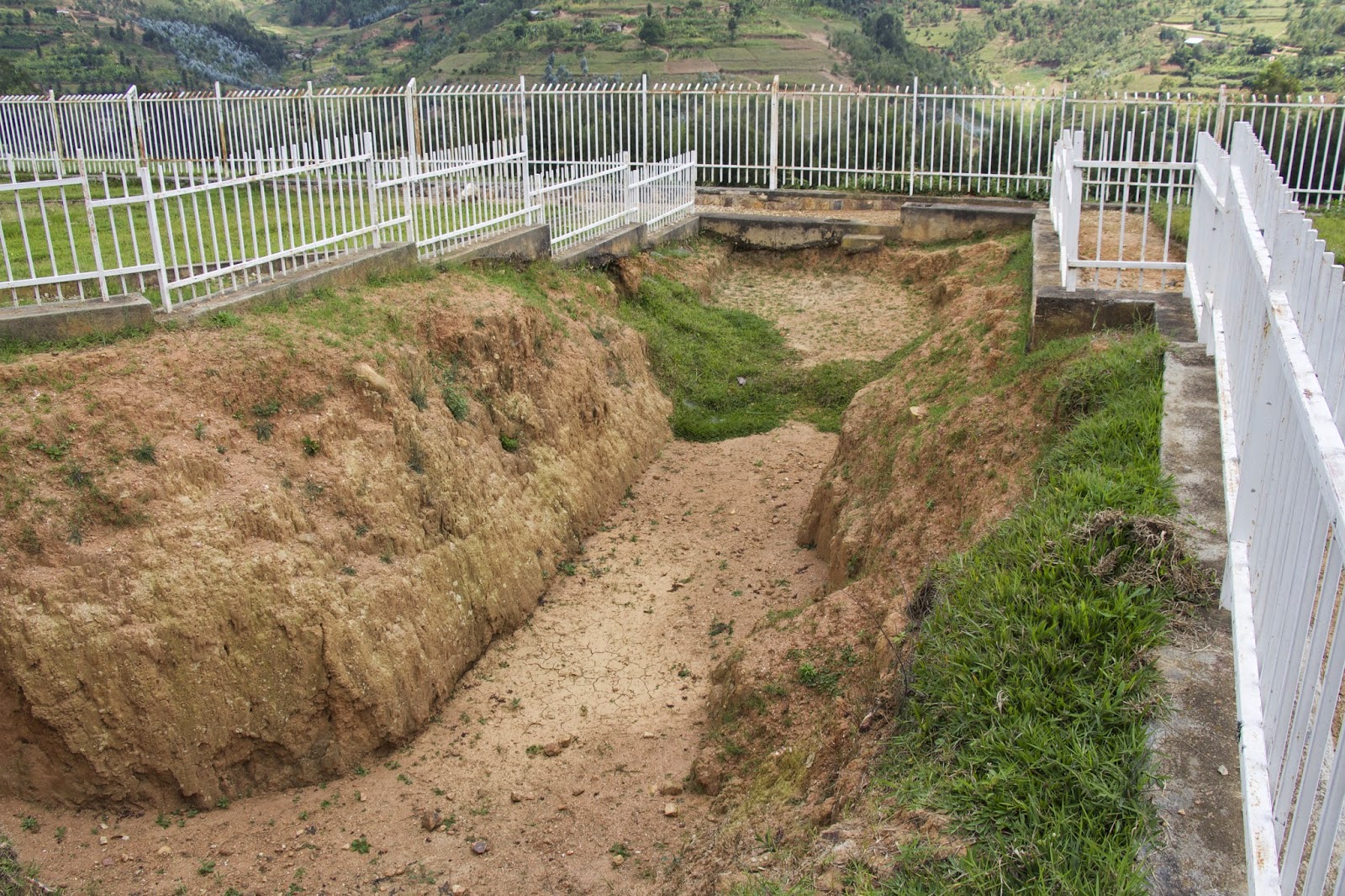 Open mass grave excavation surrounded by white fencing at the Murambi memorial, with green hills in the background