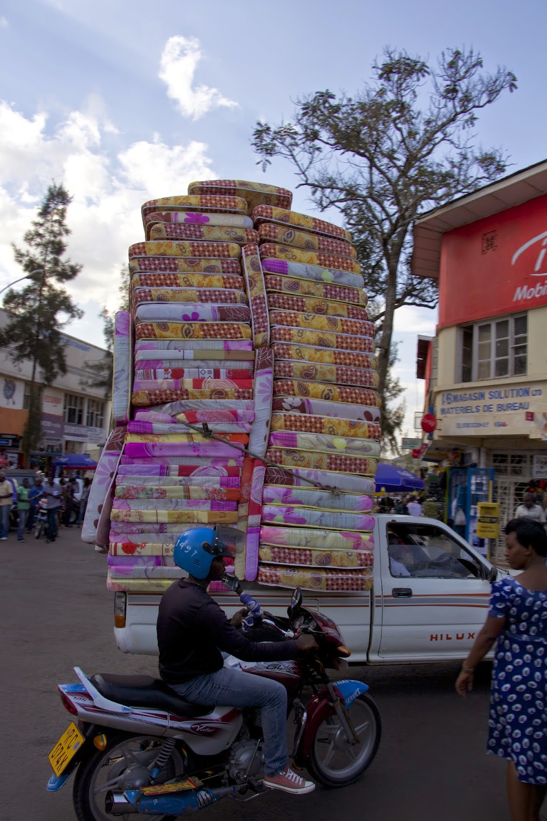 Enormous stack of colorful mattresses piled high on a pickup truck in downtown Kigali, with a motorcycle rider passing by