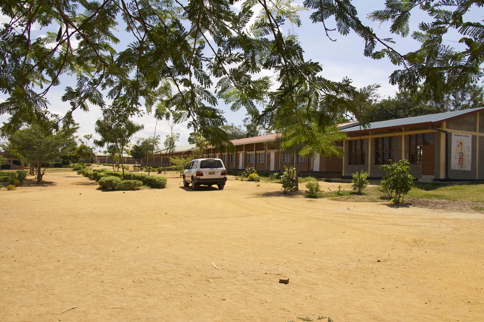 School compound with long single-story classroom buildings, sandy grounds, and trees