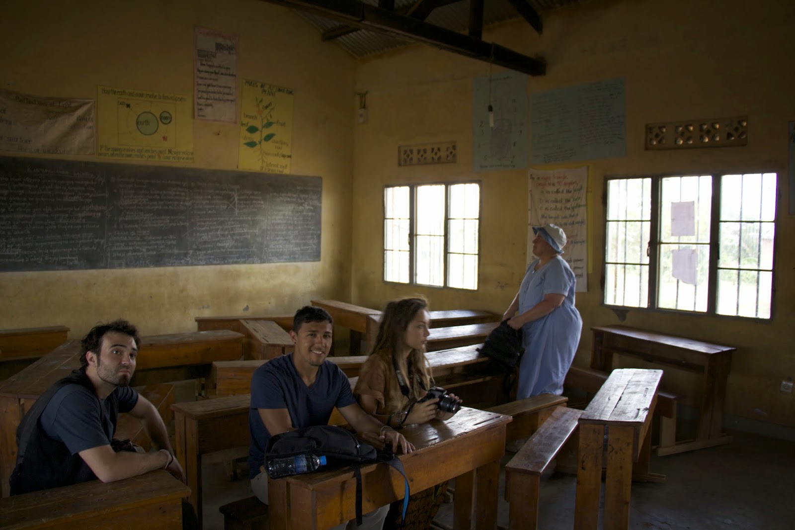Visitors sitting at wooden desks inside a village classroom with a blackboard and educational posters on the walls