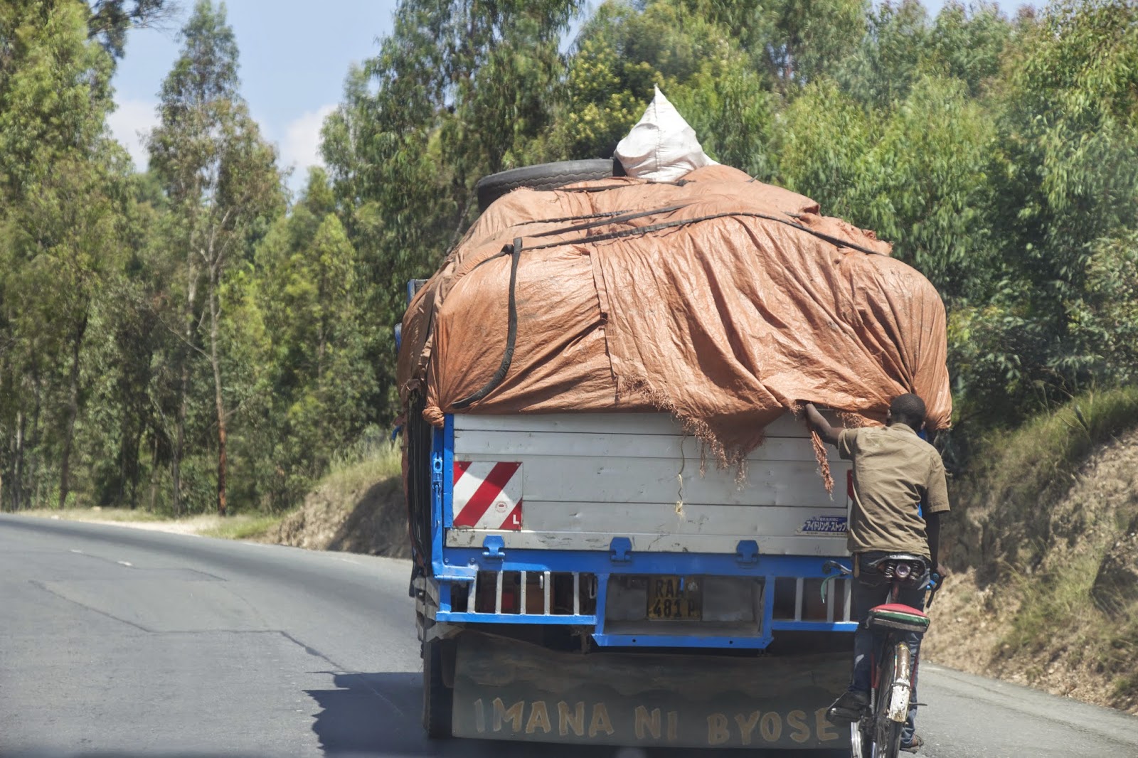 Person clinging to the back of a tarp-covered cargo truck on a Rwandan road