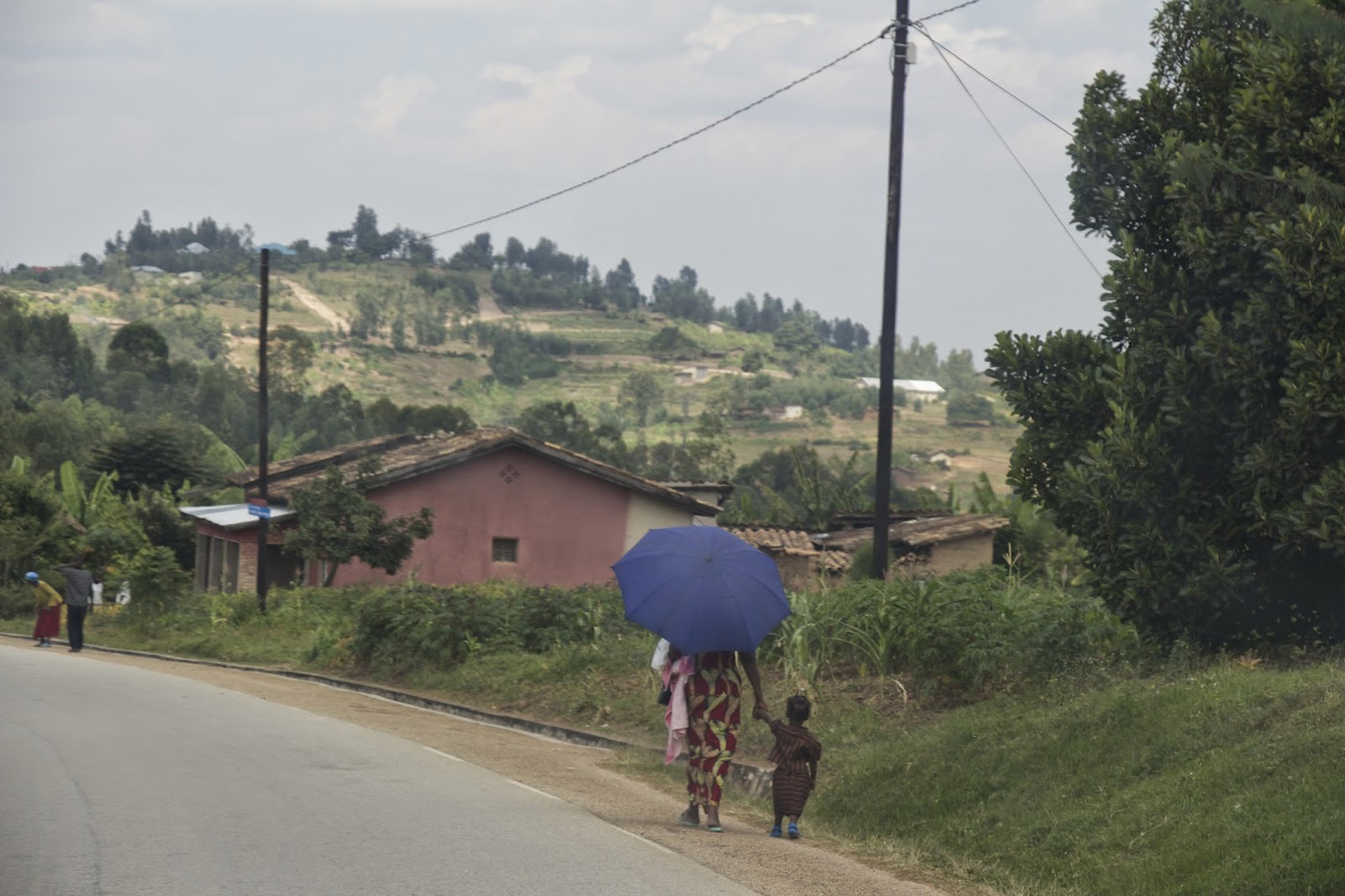 Woman carrying a blue umbrella walking along the roadside holding a small child's hand