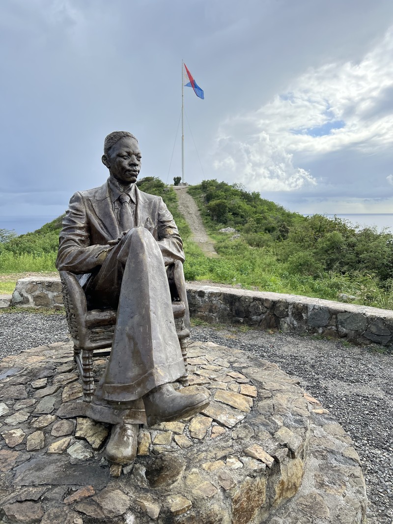 Bronze statue of a seated man on a hilltop with the Sint Maarten flag behind